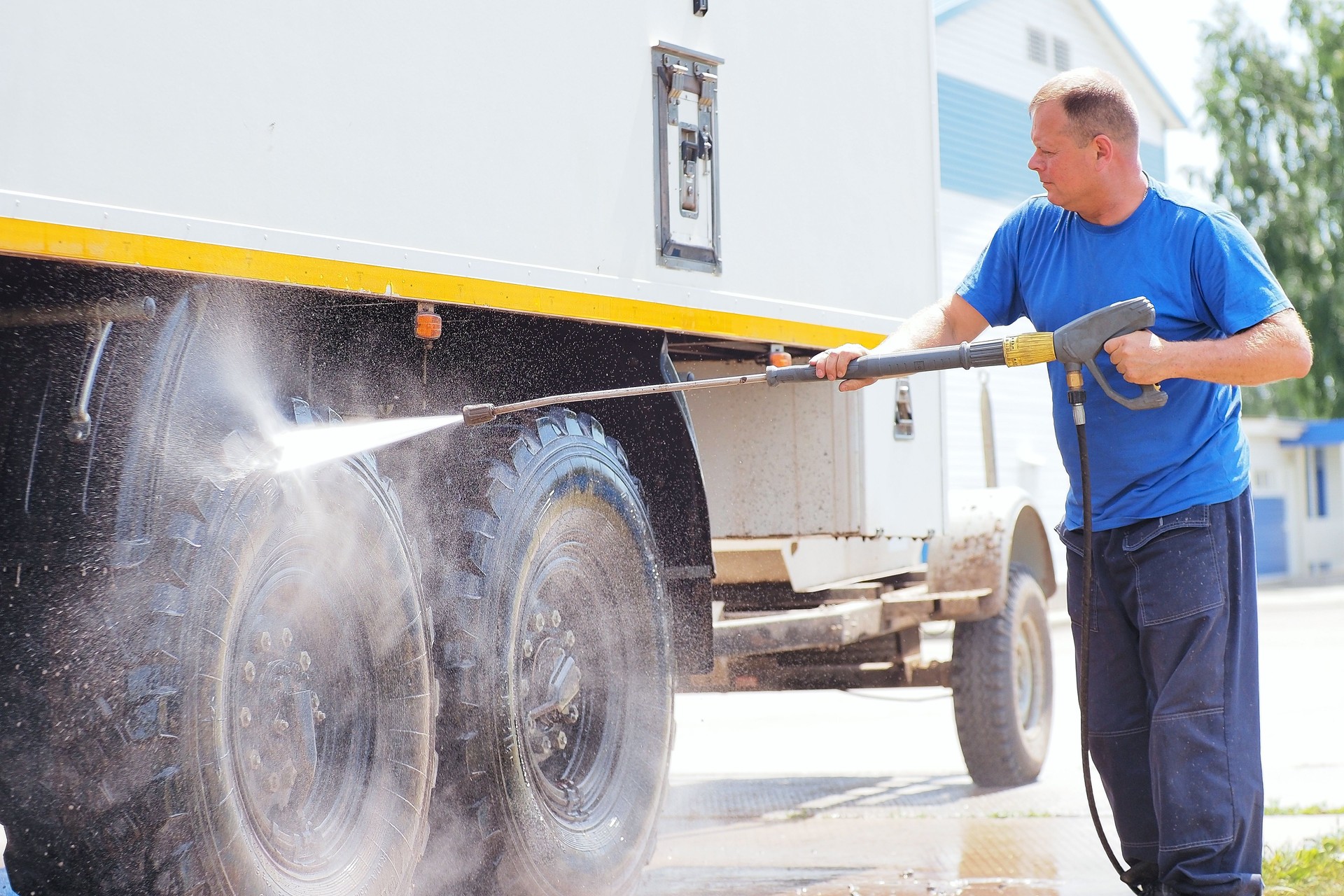 Man in overalls washes truck with water gun. Car wash for trucks. Car washer sprays car with water jet on summer day.